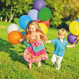 Education through movement - A girl and a boy running hand in hand with balloons across a green meadow.