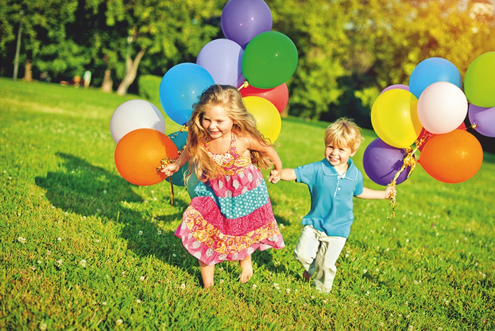 Education through movement - A girl and a boy running hand in hand with balloons across a green meadow.