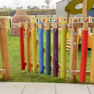 Spielplatzgestaltung – Ein Spielplatz umgeben von einem Zaun bestehend aus Latten in Form bunter Stifte.
